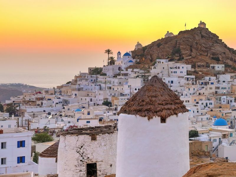 White-washed houses and an old windmill in a Greek island village at sunset, with a golden sky and a rocky hill in the background.