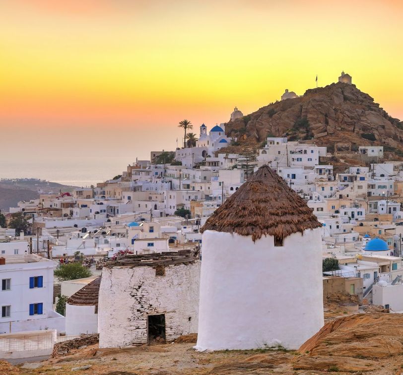 White-washed houses and an old windmill in a Greek island village at sunset, with a golden sky and a rocky hill in the background.