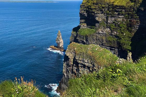 A tall, rugged cliffside covered in greenery overlooks a vast blue ocean, with a small rocky stack emerging from the water below under a clear sky.