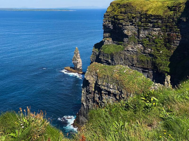 A tall, rugged cliffside covered in greenery overlooks a vast blue ocean, with a small rocky stack emerging from the water below under a clear sky.