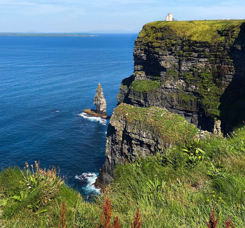 A tall, rugged cliffside covered in greenery overlooks a vast blue ocean, with a small rocky stack emerging from the water below under a clear sky.