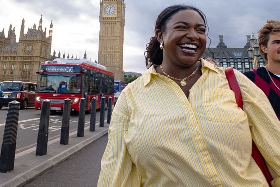 A smiling woman in a yellow striped shirt walks near Big Ben in London, with a red double-decker bus and cloudy sky in the background.