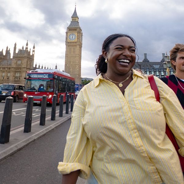 A smiling woman in a yellow striped shirt walks near Big Ben in London, with a red double-decker bus and cloudy sky in the background.