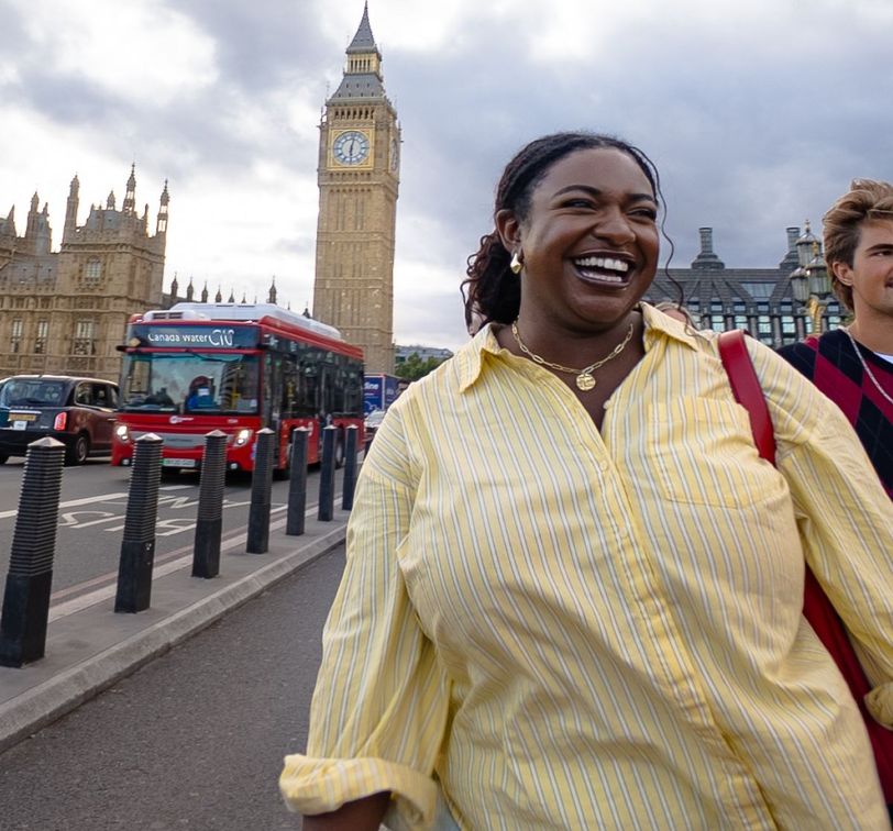A smiling woman in a yellow striped shirt walks near Big Ben in London, with a red double-decker bus and cloudy sky in the background.