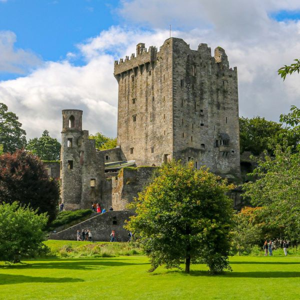 A medieval castle with tall stone walls and a central tower, surrounded by lush green lawns and trees under a partly cloudy blue sky.