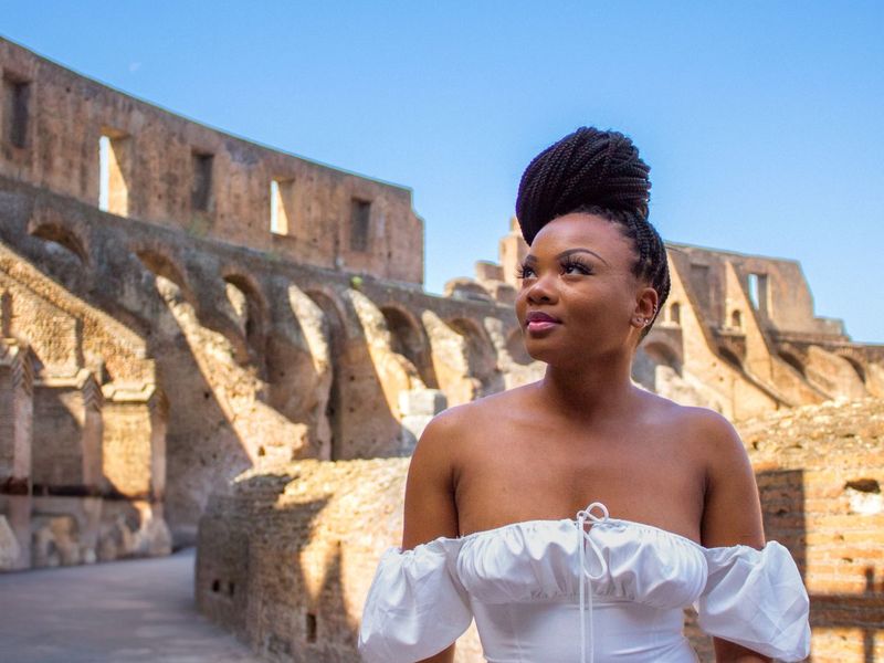A woman in a white dress stands in front of ancient stone ruins under a clear blue sky.