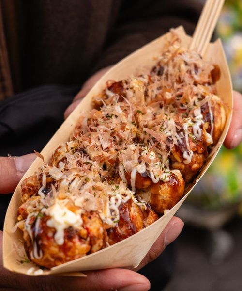 Person holding a tray of takoyaki topped with sauce, mayonnaise, and bonito flakes, garnished with herbs, using a wooden fork.