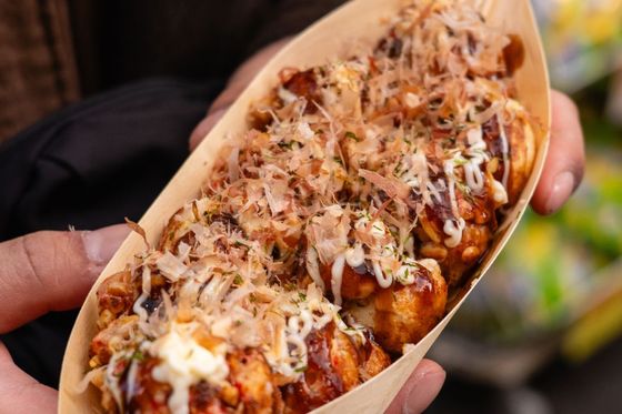 Person holding a tray of takoyaki topped with sauce, mayonnaise, and bonito flakes, garnished with herbs, using a wooden fork.