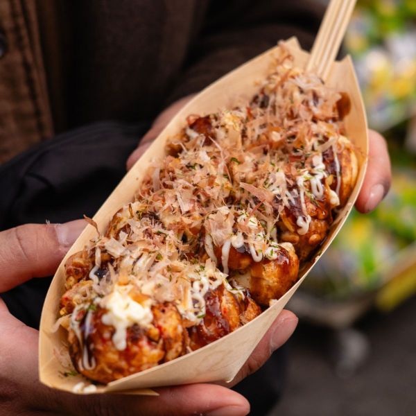 Person holding a tray of takoyaki topped with sauce, mayonnaise, and bonito flakes, garnished with herbs, using a wooden fork.
