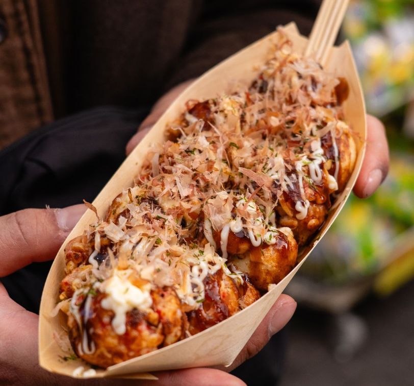 Person holding a tray of takoyaki topped with sauce, mayonnaise, and bonito flakes, garnished with herbs, using a wooden fork.