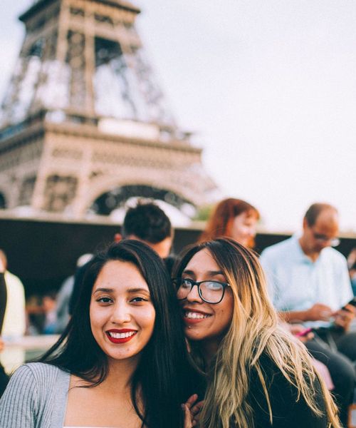 Two smiling women in front of the Eiffel Tower, with several people and the tower blurred in the background.