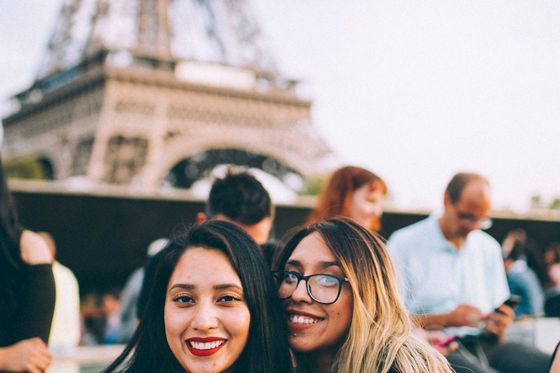 Two smiling women in front of the Eiffel Tower, with several people and the tower blurred in the background.