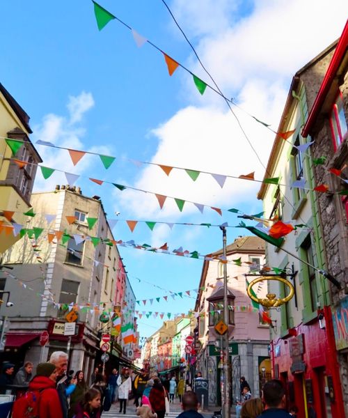 A lively street scene with colorful buildings, festive bunting, and people walking. The sky is blue with a few clouds, creating a cheerful atmosphere.