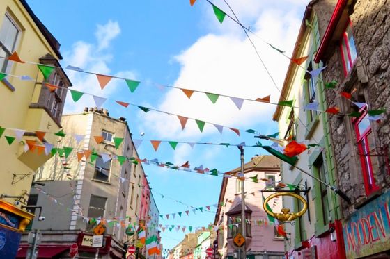 A lively street scene with colorful buildings, festive bunting, and people walking. The sky is blue with a few clouds, creating a cheerful atmosphere.