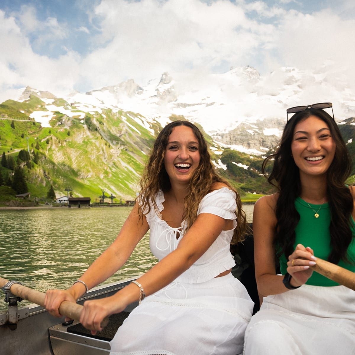 Two women smiling while rowing a boat on a lake, surrounded by snow-capped mountains and lush greenery under a partly cloudy sky.