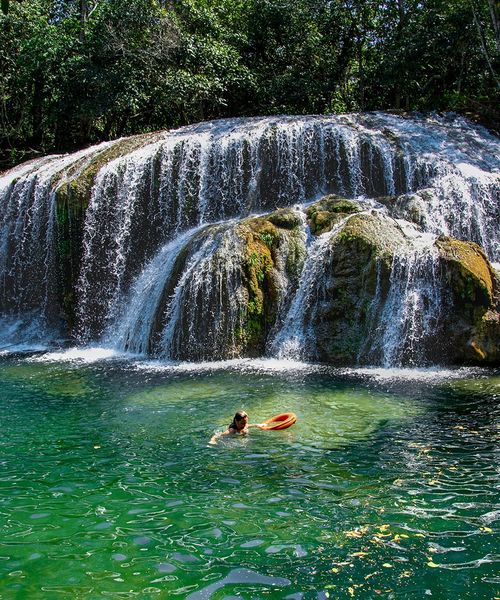 Person swimming near a cascading waterfall in a green forest setting, holding an orange flotation ring in clear, emerald water.