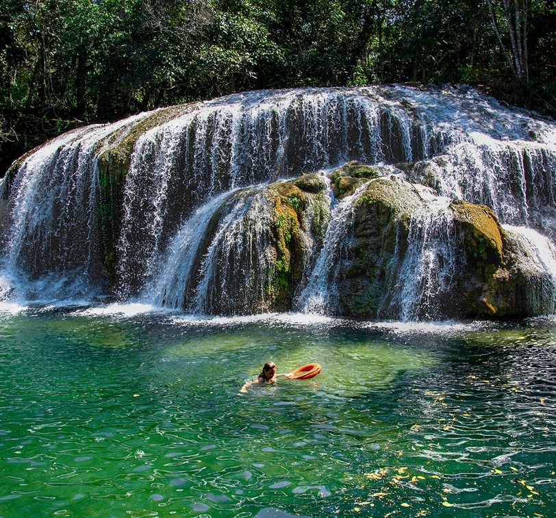 Person swimming near a cascading waterfall in a green forest setting, holding an orange flotation ring in clear, emerald water.