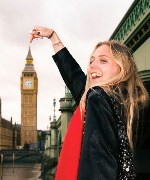 Smiling woman points at the iconic clock tower in London, standing near a bridge with cloudy skies in the background.