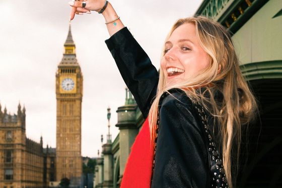 Smiling woman points at the iconic clock tower in London, standing near a bridge with cloudy skies in the background.