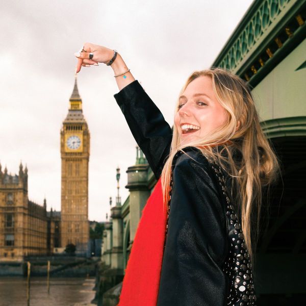Smiling woman points at the iconic clock tower in London, standing near a bridge with cloudy skies in the background.