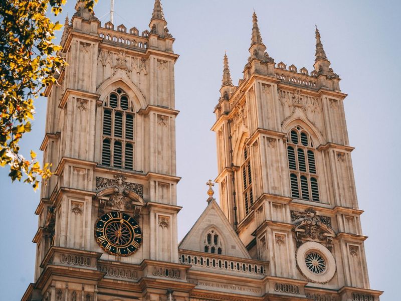 A close-up of the two towers of Westminster Abbey, with intricate Gothic architecture and a blue sky background framed by tree branches.