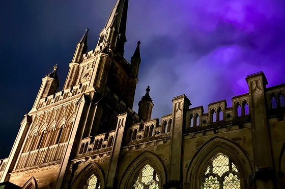 Gothic cathedral with illuminated stained glass windows and a dramatic purple sky at dusk.