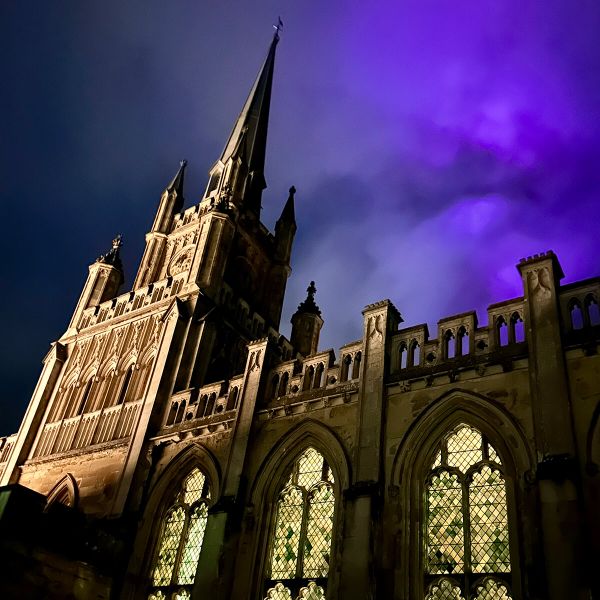 Gothic cathedral with illuminated stained glass windows and a dramatic purple sky at dusk.