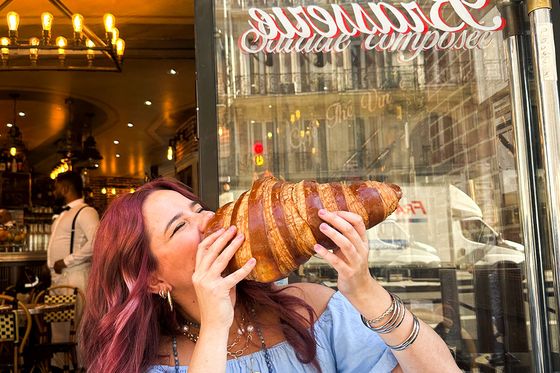 A woman with red hair joyfully holds and pretends to bite a giant croissant outside a café with elegant decor.