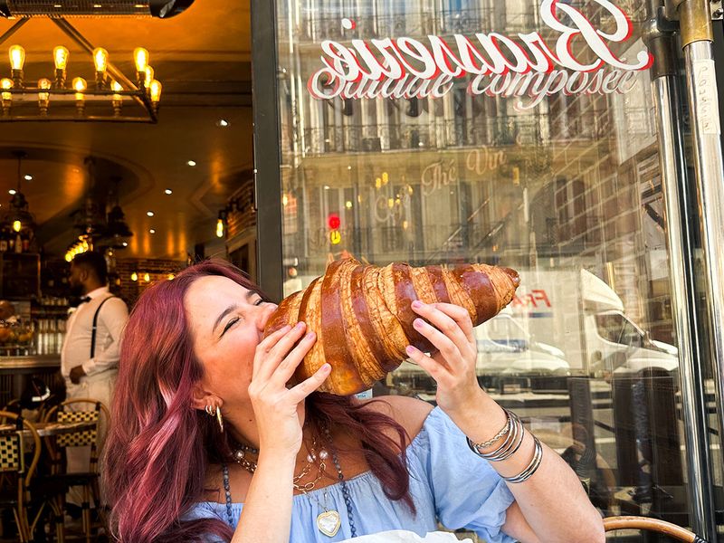 Person playfully holding a giant croissant in front of a cafe in Paris. Bright street reflections in the window add a lively atmosphere.