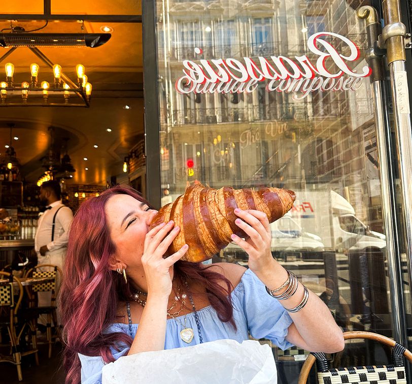 Person playfully holding a giant croissant in front of a cafe in Paris. Bright street reflections in the window add a lively atmosphere.