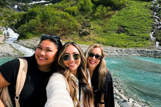 Three women smiling and taking a selfie by a turquoise mountain stream, with lush green hills and a snow-capped peak in the background.