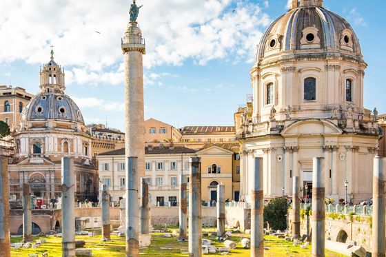 Ancient Roman ruins with a tall column and two domed churches in the background, set against a bright blue sky with scattered clouds.