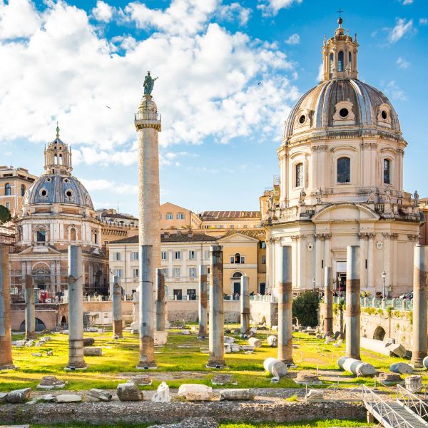Ancient Roman ruins with a tall column and two domed churches in the background, set against a bright blue sky with scattered clouds.