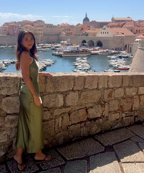 Woman in a green dress stands by a stone wall with a scenic view of boats in a historic harbor and old town with terracotta roofs.