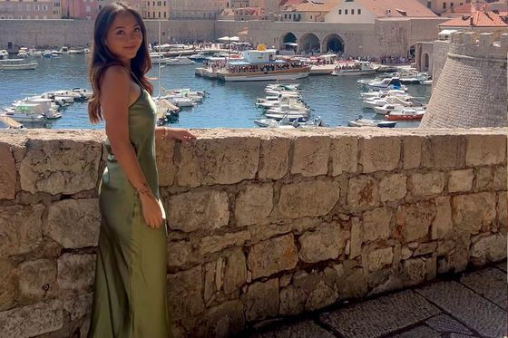 Woman in a green dress stands by a stone wall with a scenic view of boats in a historic harbor and old town with terracotta roofs.