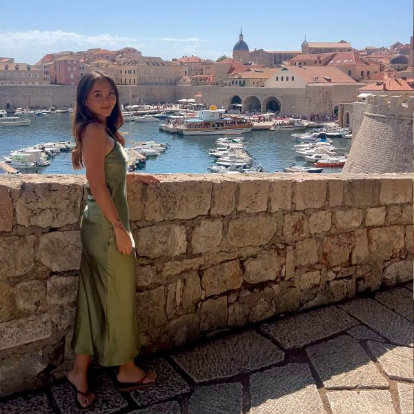 Woman in a green dress stands by a stone wall with a scenic view of boats in a historic harbor and old town with terracotta roofs.