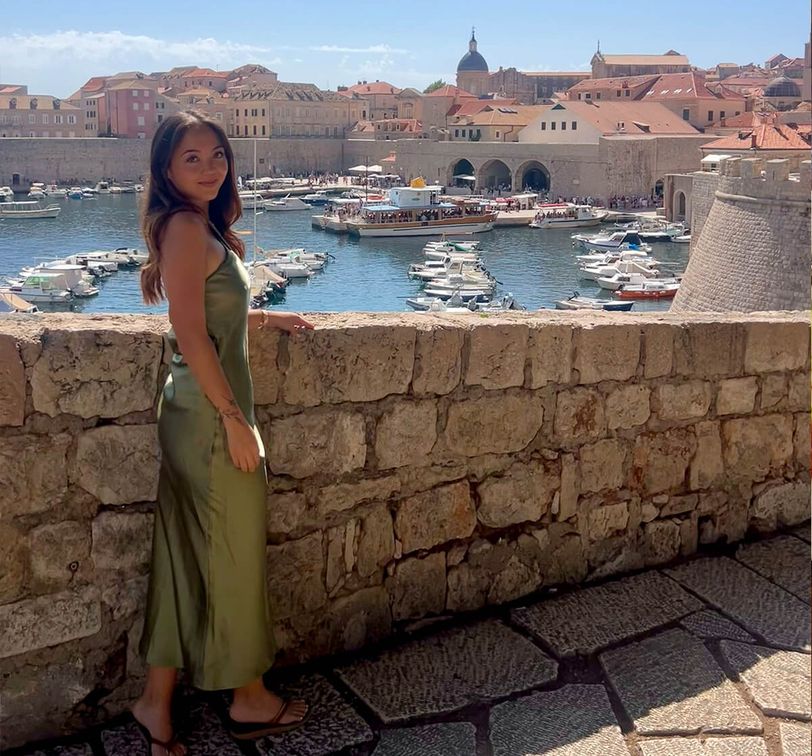 Woman in a green dress stands by a stone wall with a scenic view of boats in a historic harbor and old town with terracotta roofs.