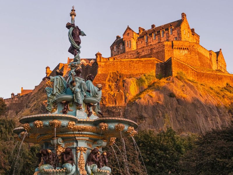 A historic castle on a hilltop is bathed in golden sunlight with an ornate, turquoise fountain in the foreground, surrounded by greenery.