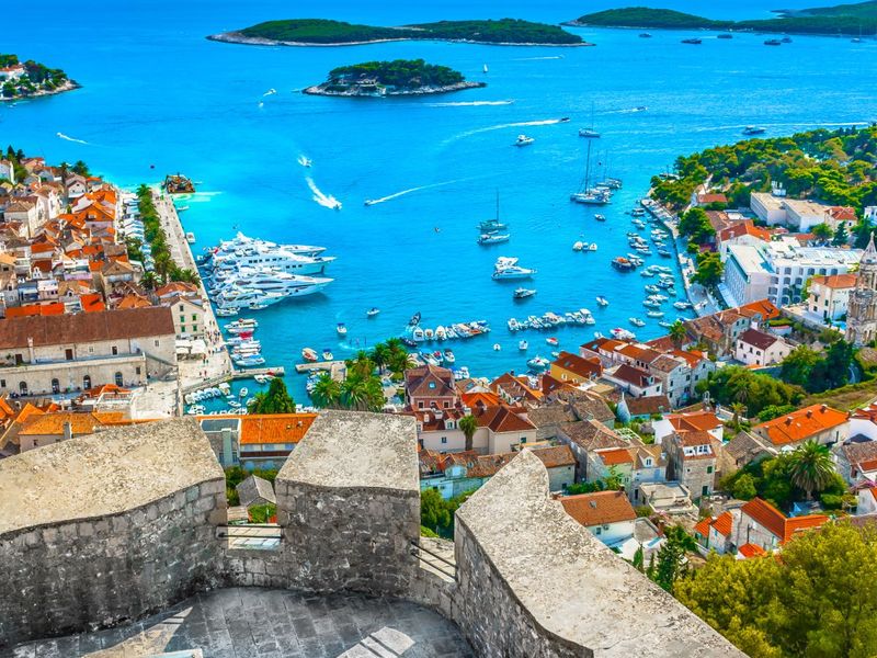 Scenic view of a coastal town with red-roofed buildings, sailing boats, and lush islands in a vibrant blue sea, seen from a high stone terrace.