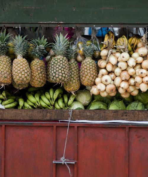 A variety of fruits and vegetables on display at a market stall, featuring pineapples, bananas, onions, and lettuce against a wooden and red backdrop.