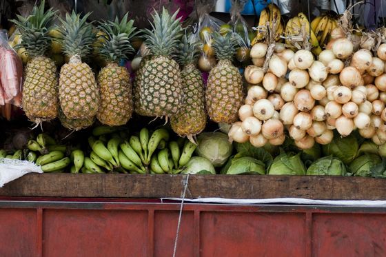 A variety of fruits and vegetables on display at a market stall, featuring pineapples, bananas, onions, and lettuce against a wooden and red backdrop.