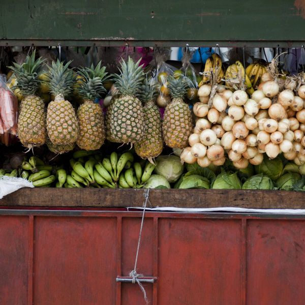 A variety of fruits and vegetables on display at a market stall, featuring pineapples, bananas, onions, and lettuce against a wooden and red backdrop.