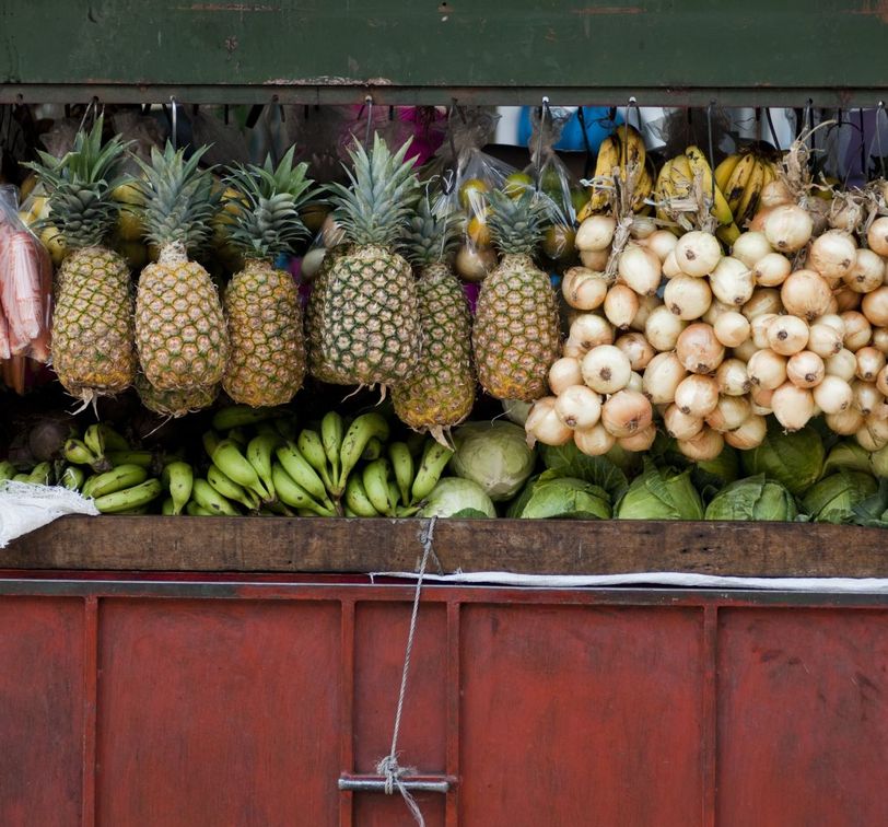 A variety of fruits and vegetables on display at a market stall, featuring pineapples, bananas, onions, and lettuce against a wooden and red backdrop.