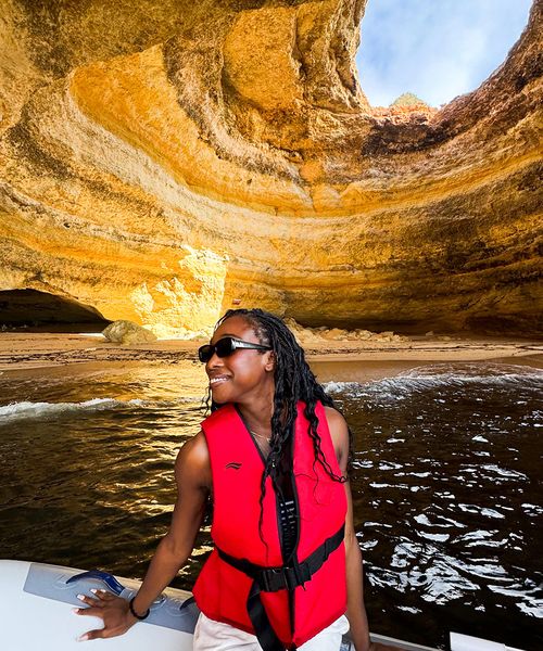 A person in a red life jacket smiles while standing on a boat inside a sunlit sea cave with layered rock formations.