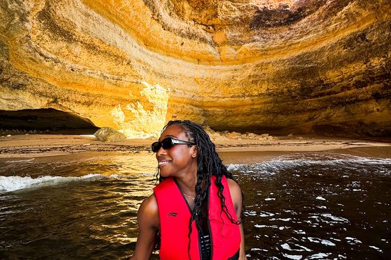 A person in a red life jacket smiles while standing on a boat inside a sunlit sea cave with layered rock formations.