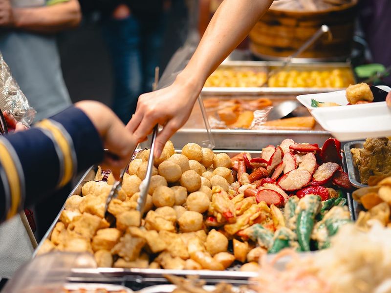 Street food vendor serving various fried snacks, including tofu and vegetables, with tongs at a bustling market stall.