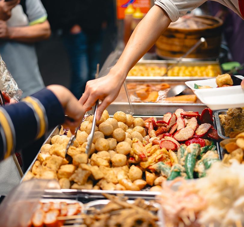 Street food vendor serving various fried snacks, including tofu and vegetables, with tongs at a bustling market stall.