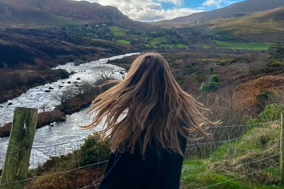 Person with long hair stands near a railing, overlooking a river and green hills under a partly cloudy sky.