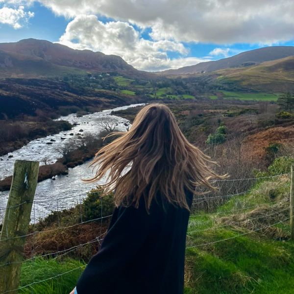 Person with long hair stands near a railing, overlooking a river and green hills under a partly cloudy sky.