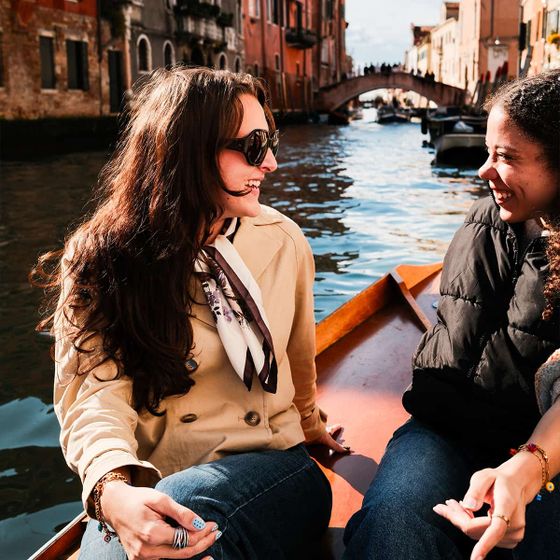 Two women smiling and conversing on a boat ride through a canal, with historic buildings and a bridge in the background.